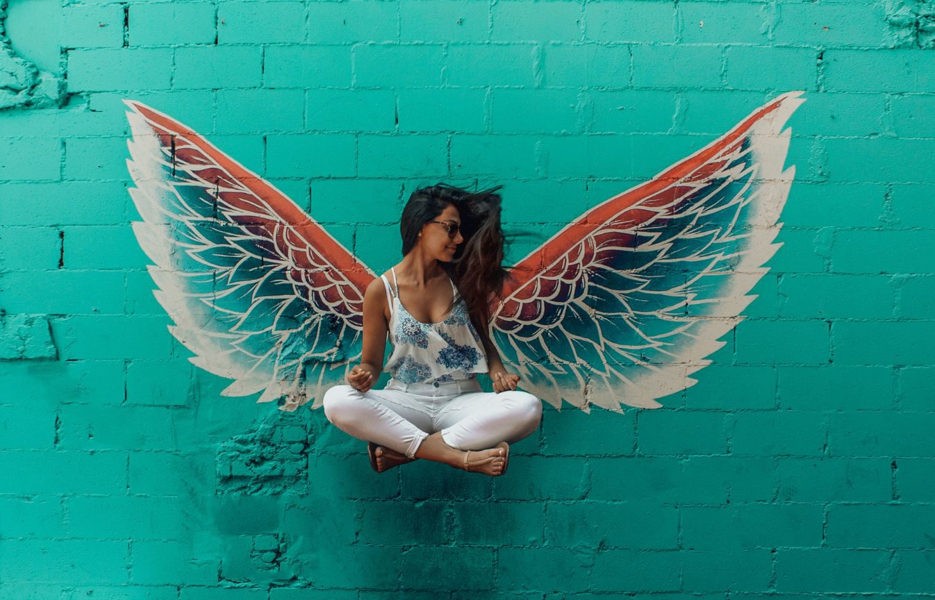 3 Phasen der Selbstfindung, Phase 3: Entfalten, Frau sitzt im Schneidersitz vor einer grünen Mauer mit großen, gemalten Flügeln 3 Phasen der Selbstfindung, Phase 3: Entfalten, Frau sitzt im Schneidersitz vor einer grünen Mauer mit großen, gemalten Flügeln