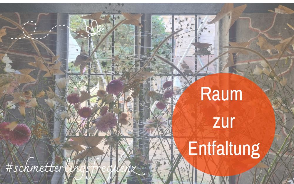 Ein antikes Fenster mit Sprossen, davor ein filigran arrangiertes Blumenkunstwerk mit Papierschmetterlingen. Text: Raum zur Entfaltung